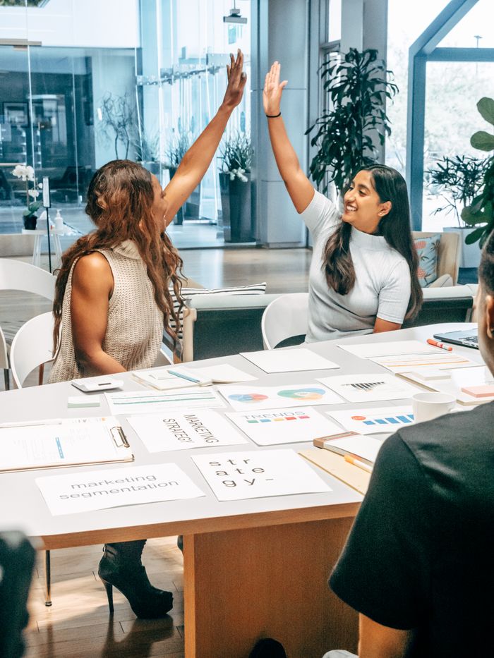 Two women in an office share a high five, symbolizing successful teamwork and cooperation.
